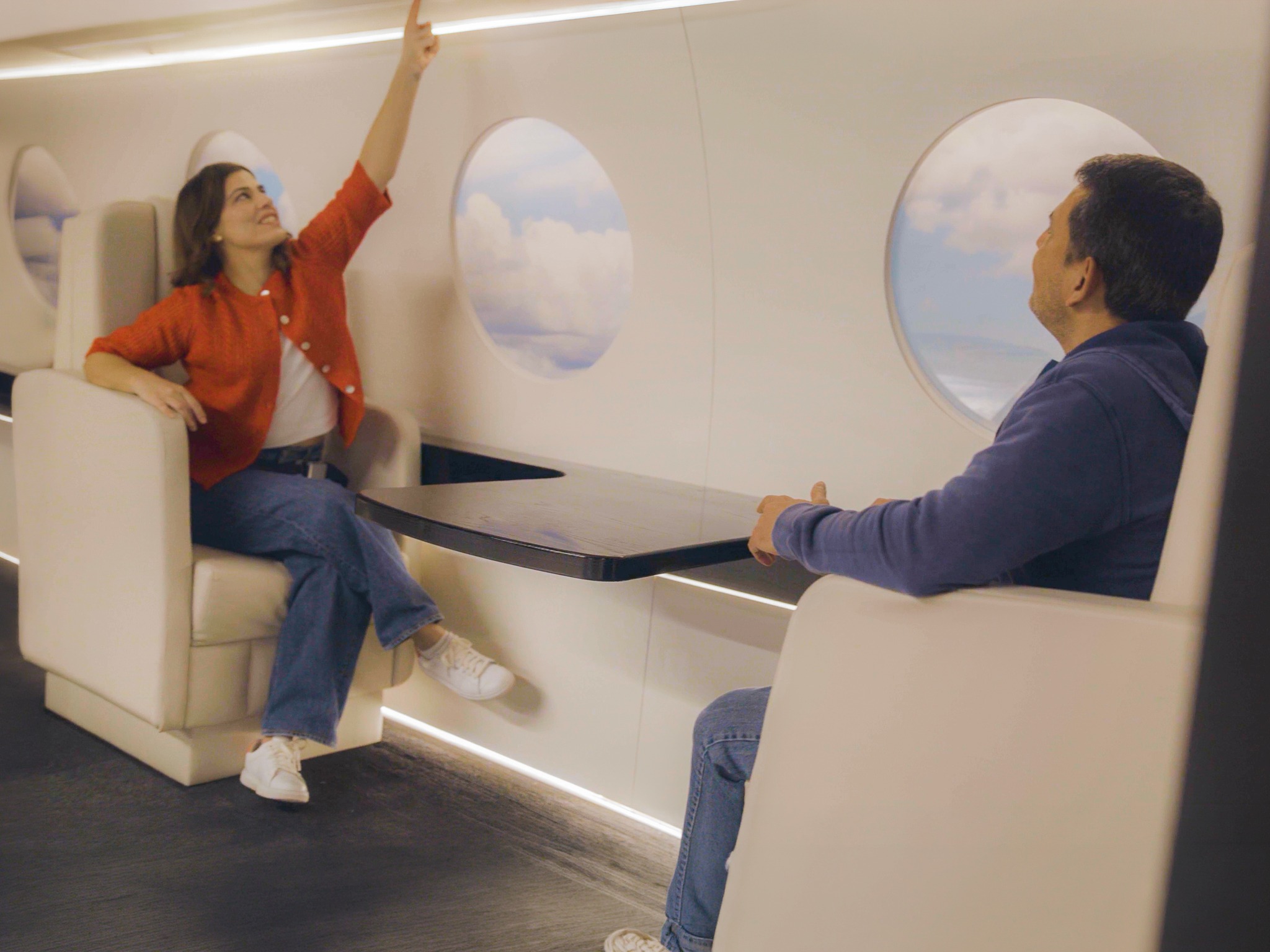 Couple at a sleek table in Lintshiwe's private jet exhibit, with painted clouds visible in the round windows behind them.