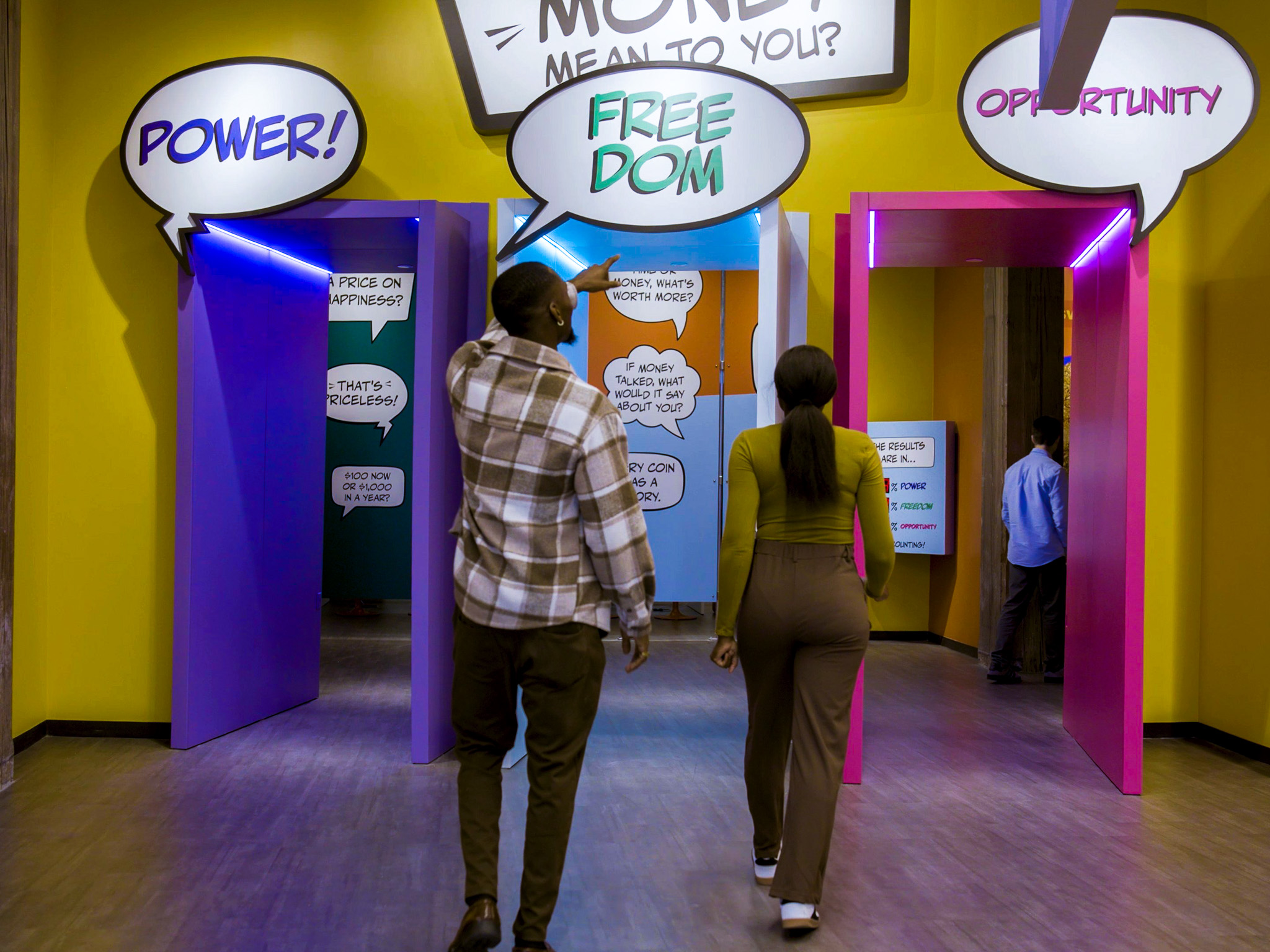 Couple walks through Lintshiwe's bright corridor past a comic-style speech-bubble exhibit asking what money means.