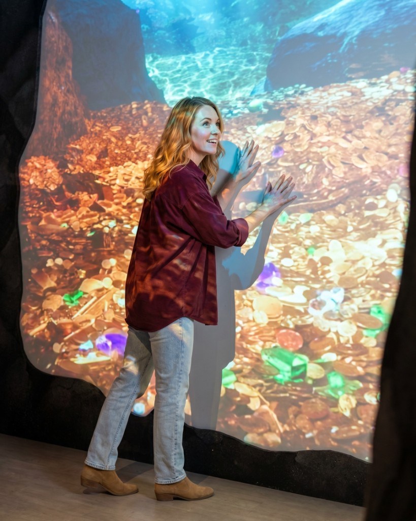 Woman presses her hands against Lintshiwe's glowing wall of gold coins and gems at the Hoard of Gold exhibit, smiling in wonder.